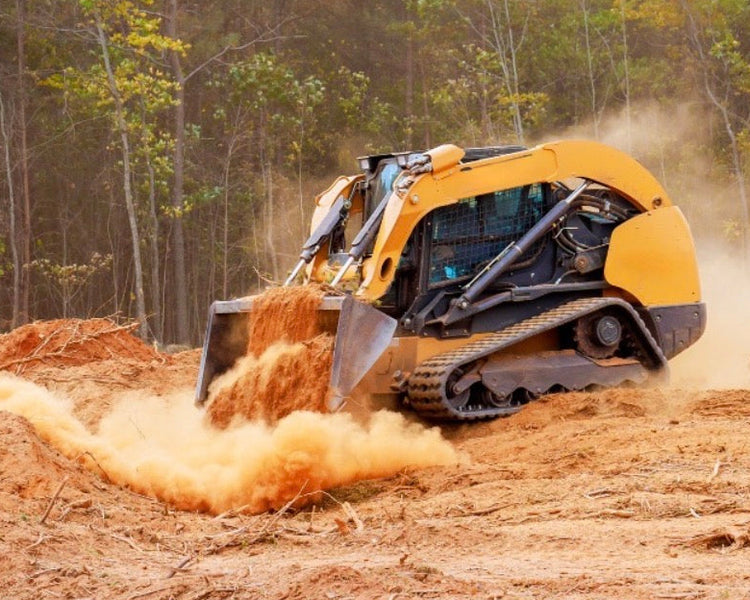 A yellow compact skid steer loader is shown in a real outdoor construction environment, pushing dirt and mulch with a cloud of dust behind it, with a forest in the background.