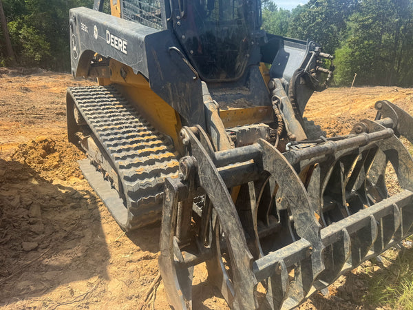 A John Deere skid steer loader with a large front grapple attachment, parked on a dirt construction site with trees in the background.