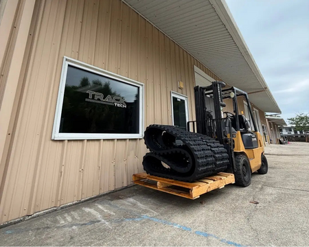 Yellow forklift with large black rubber tracks on a wooden pallet in front of a  TrackTECH rubber tracks, Destin, Florida office.